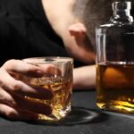 A person with their head down, holding a glass of whiskey next to a bottle