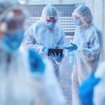Healthcare workers in protective gear discussing information on a tablet outdoors