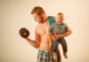 A muscular man lifting a dumbbell while holding a smiling baby