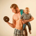 A muscular man lifting a dumbbell while holding a smiling baby