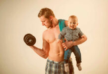A muscular man lifting a dumbbell while holding a smiling baby