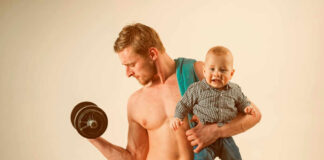 A muscular man lifting a dumbbell while holding a smiling baby