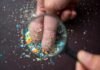 A hand holding a magnifying glass over fingers covered in colorful particles