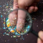 A hand holding a magnifying glass over fingers covered in colorful particles