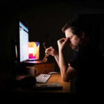 A man sitting at a desk with a computer, holding glasses and looking stressed