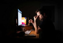 A man sitting at a desk with a computer, holding glasses and looking stressed