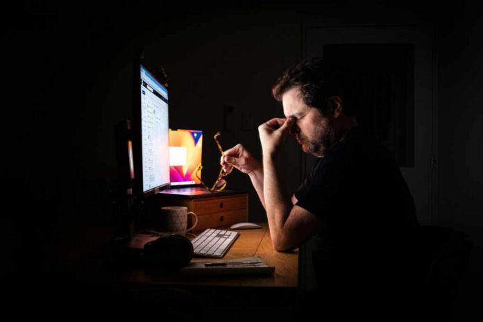 A man sitting at a desk with a computer, holding glasses and looking stressed