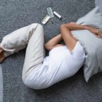 Person lying on a bed with a pillow over their head, surrounded by medication
