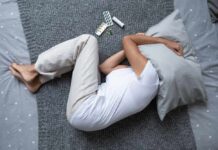 Person lying on a bed with a pillow over their head, surrounded by medication