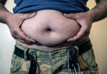 Hidden Fat-Burning Switch Found Man holding his belly with both hands, wearing a blue shirt and camouflage pants