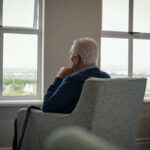Elderly man sitting in a chair, looking out a window with a thoughtful expression