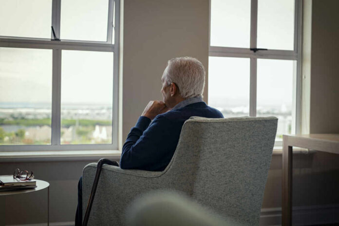 Elderly man sitting in a chair, looking out a window with a thoughtful expression