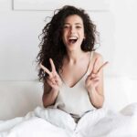 Smiling woman on bed showing double peace signs