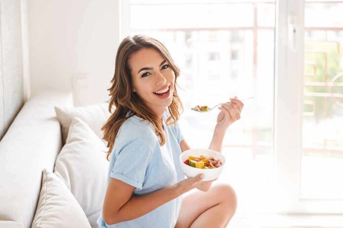 shutterstock_1136119265 Woman sitting on a couch eating a bowl of fruit salad