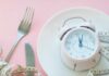 This Eating Pattern Was Linked To Better Memory In Seniors Pink alarm clock on a white plate with fork, knife, and measuring tape
