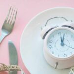 Pink alarm clock on a white plate with fork, knife, and measuring tape
