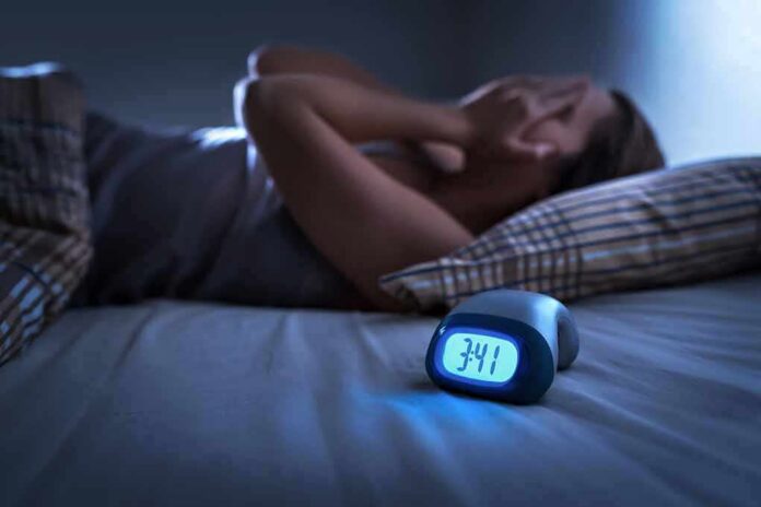 shutterstock_1299304069 Person lying awake in bed beside a glowing digital alarm clock
