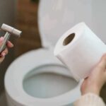 Hands holding a bidet sprayer and toilet paper near an open toilet