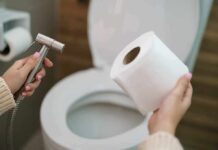 Hands holding a bidet sprayer and toilet paper near an open toilet
