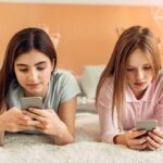 Two girls lying on a bed using smartphones