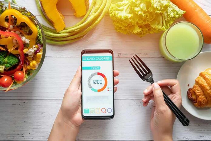 shutterstock_1465238459 hands holding phone with calorie tracker beside salad and pastry