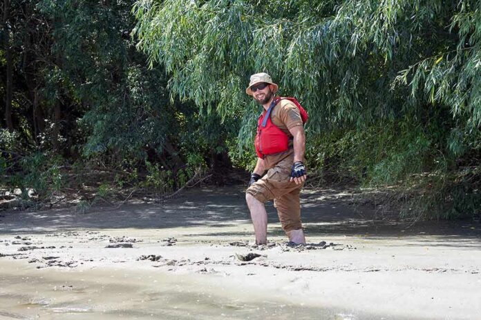 shutterstock_1629856273 man in red life vest walking through muddy riverbank