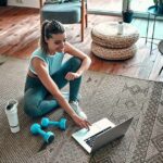 Woman in workout clothes using a laptop on a living room rug