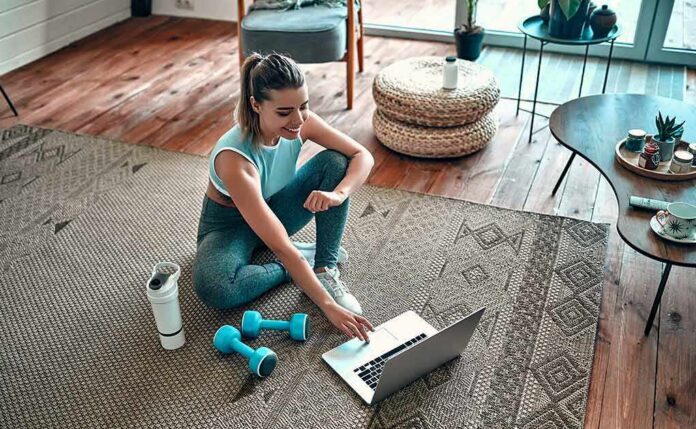 shutterstock_1660419799 Woman in workout clothes using a laptop on a living room rug