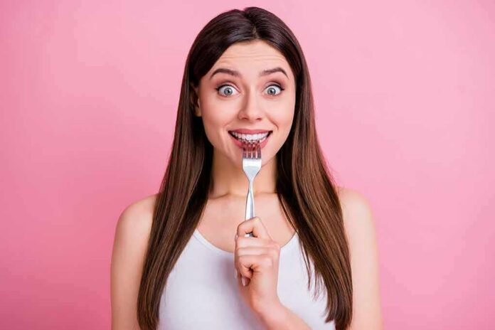 shutterstock_1740348350 Woman holding a fork to her teeth against a pink background
