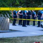 police officers stand behind caution tape at a park scene