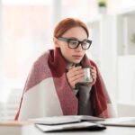 Woman in glasses wrapped in a blanket holding a mug at a laptop