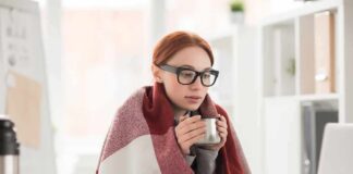 Woman in glasses wrapped in a blanket holding a mug at a laptop