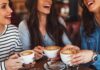 Three friends laughing while holding cups of cappuccino at a café table