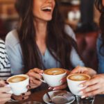 Three friends laughing while holding cups of cappuccino at a café table