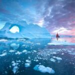 Iceberg with arch and red-sailed boat at colorful sunset