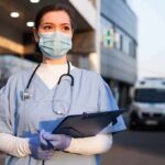 Healthcare worker in scrubs wearing mask and gloves holding a clipboard