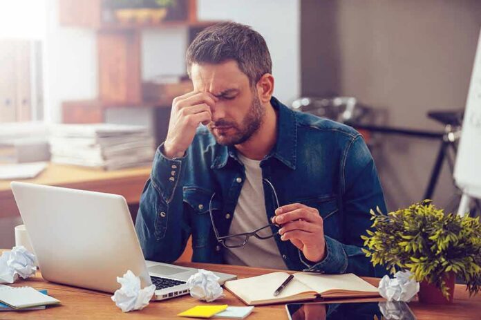 Man rubbing his eyes at a desk with a laptop and crumpled papers